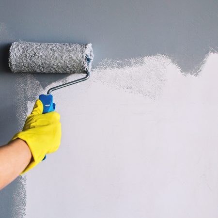 Hand in yellow glove painting wall in gray color with a roller brush.